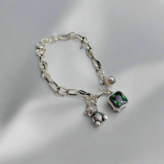 Close-up view of the Radiant Teddy Charm Bracelet featuring a silver chain, teddy bear charm, and green gemstone charm displayed on a white background.